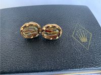 Pair of oval cuff links with faceted art glass centers featuring multi-color horizontal stripes, set in textured gold-tone mounts, placed on black leather with gold print.