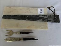 Full view of rectangular white marble cheeseboard with attached narrow black marble handle and brown leather strap, alongside two serving utensils with wooden handles.