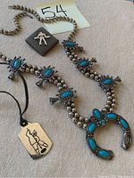 Photo of silver Inuit pendant dog tag with engraved figure on black cord, Navajo style silver and turquoise squash blossom necklace and matching bracelet, and square leather brooch with bead figure.