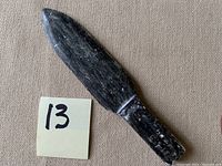 Full view of the primitive dark stone spearhead on beige fabric with size note '13'