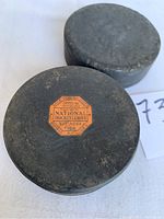 Photo showing two older black hockey pucks, one with an orange label on top with NHL Art Ross Tyler text, the other plain black rubber puck.