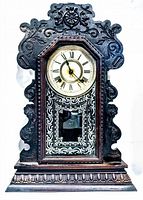 Front view showing wooden clock case with intricate carvings, round clock face with Roman numerals, and palm leaf stenciled design on lower glass.