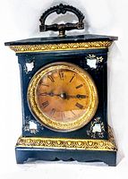 Front view showing black rectangular case with ornate gold-tone trim and mother of pearl accents around the clock face. The clock face is round with gold-tone bezel and Roman numerals.