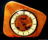 Front close-up image of the clock showing the round face, cream dial with black Arabic numerals, and light wood asymmetrical case.