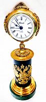 Front angled view of vintage quartz table clock with gold-toned round face, black Roman numerals, and gold ornamental details on black cylindrical body above green stone base.