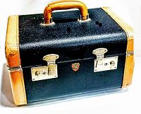 Closed vintage vanity case showing black textured exterior, tan leather corners, silver metal clasps, handle, and McBrine Baggage logo.