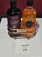 Front view of Beekman Fig Leaf Goat Milk and Awakening Honeyed Grapefruit hand and body wash bottles in metal holder, on a white surface with lot number card 25.