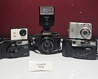 Front view of all six cameras including Canon T50 with flash, Yashica MG-2, Nikon One Touch 100, Nikon Coolpix L4, generic 720P camera, and silver Nikon digital camera arranged on white surface.