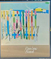 Front view of the framed Dan Poole Rainbow Beach print showing colorful vertical fence posts on the beach with houses in the background.