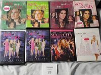 Photo of eight Sex And The City DVD sets on a white surface, showing different season and volume cases, some sealed and some open, with visible HBO branding.