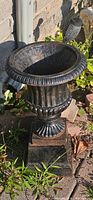 Photo of small cast iron planter in garden setting, showing the urn-shaped planter with fluted details and square pedestal base, with signs of weathering.