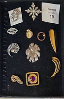Top view of a black tray holding 11 unsigned vintage brooch pins in various designs showing silver-tone and gold-tone metals with rhinestone embellishments.
