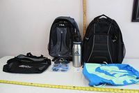 Overall shot of backpacks, bags, sunglasses and water bottle assembled on table with measuring tape for size reference.