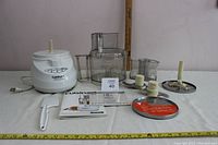 Full lot with motor base, bowls, blades, discs, spatula, and manuals arranged on a white surface against a plain wall, showing all components included.