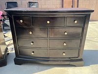 Photo of dark cherry wood dresser with 9 drawers shown from the front outdoors, with metal round drawer knobs and a bowed front panel.