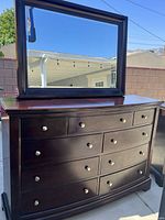 Photo showing a dark wood dresser with nine drawers and round metal knobs, topped with a rectangular attachable mirror with a matching dark wood frame.