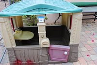 Front side view showing purple door, beige walls with lower textured detail mimicking stone, yellow sink with faucet inside, and red window shutters.