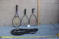 Three rackets (tennis, squash, racquetball) displayed upright with ruler for scale on gray background, including a Head carrying case laid in front