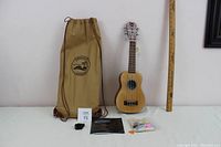 Ukulele with carrying bag, tuner, instruction booklet, picks, and strings arranged with measuring stick for size reference.