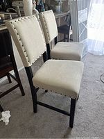 Photo showing two cream fabric and wood bar stools with backs, placed indoors on carpeted floor next to kitchen counter.