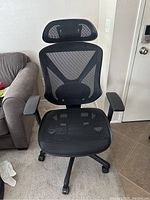 Front view of black mesh ergonomic office chair with headrest and armrests positioned upright indoors on tiled floor near couch.