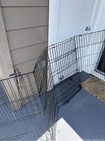 Black metal dog fencing panels arranged outside on a porch or balcony surface. Panels shown bent to configure an enclosure with an entrance panel visible.