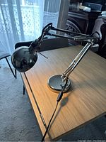 Side view of the adjustable IKEA metal desk lamp placed on wooden table near window, showing chrome finish and the lamp arm with springs.