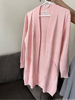 Front view of pink open-top sweater hanging on hanger, showing long sleeves, open front, pockets and ribbed hem and cuffs.