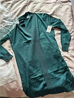 Full view of the teal women's sweater laid flat, showing ribbed cuffs and front pockets, with original plastic packaging included.