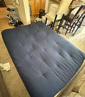 Queen-sized blue fabric futon mattress lying flat on carpeted floor in living room setting with kitchen and chairs visible in background. Mattress shows tufted details in fabric and no visible damage.