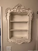 Frontal view showing the ornate carved frame and two shelves inside. Worn white paint finish visible.