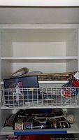 Photo of shelf with wire basket containing several rolled magazines, baseball pin items, framed baseball photo, and baseball on a lower shelf with magazines underneath.
