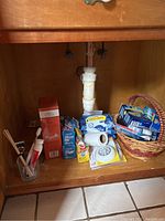 Photo showing various bath and cleaning supplies under cabinet including toothbrushes, orange boxed tissue bar, waste bags, baby wipes, small toilet paper roll, and battery operated light.