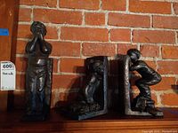 Three bronze bookends with child figures on an L-shaped base, placed against a brick wall, side view in low lighting.