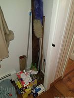 Photo of corner with various cleaning tools including purple dust mop, beige duster and green dustpan.
