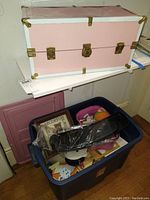 Pink metal doll trunk stacked on top of blue plastic tote filled with toys and dollhouse furniture.