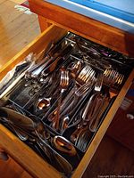 Drawer containing multiple metal organizer trays filled with mixed stainless steel flatware including spoons, forks, knives with wooden handles and cake server
