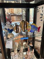Wide view showing all four glass figurines on a glass shelf in a display case: three fish figurines and one bird figurine