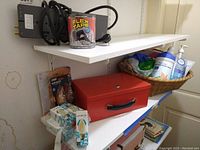 View of shelves showing light bulbs, red metal safe box, basket with sanitizing wipes, and a roll of Flex Tape with Belkin power outlet.