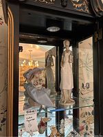 Two Lladro figurines displayed inside a glass cabinet, showing one seated girl with a hat holding flowers and one standing girl in an apron.