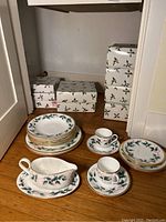 Full set showing stacks of dinner plates, smaller plates, cups and saucers, and gravy boat with underplate. Boxes with holly print are stacked in background.