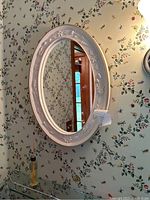 Front view of oval mirror with white embossed floral frame hanging on floral wallpapered wall.