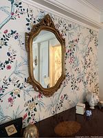 Wall mirror hanging on wallpapered wall with floral and botanical print, showing the full view of the mirror and its ornate bronze-toned frame.