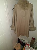 Beige wool poncho with large fur collar and fur cuffs hanging on door.