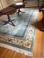 Full view of the rectangular rug under furniture showing light blue/green central area with floral patterns and cream colored border with scrolling motifs and fringe ends.