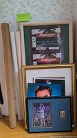 Framed Budweiser ticket stub images and stacked posters with some rolled, leaning against wall