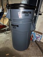 One stacked Rubbermaid Brute trash bin set showing gray bin with black lid, commercial labeling, side handles, lightly used condition. Located in garage with surrounding boxes and items.