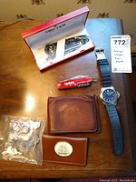 Overview of lot showing pen box, red multitool, black strap wristwatch, two brown wallets, and sealed bag of locks and keys.