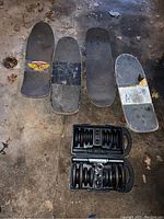 Four skateboard decks lined up above open dumbbell case on concrete floor