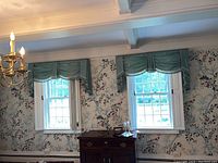 View of two windows each with a green scalloped fabric valance with tassel fringe along the bottom edges. Light blue and green floral wallpaper in background, part of interior room setting.